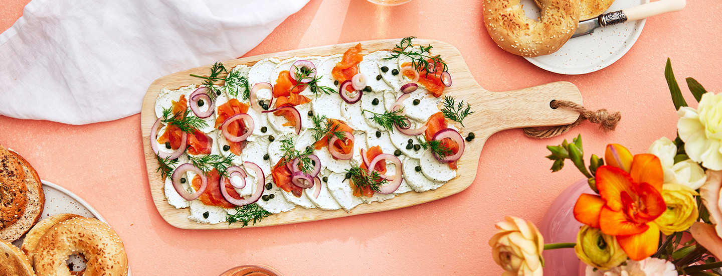 Tuorejuustoa puisella laudalla. Tuorejuuston päälle on aseteltu kylmäsavulohen viipaleita, punasipulin siivuja, kapriksia ja tilliä. Puisen laudan ympärillä on bageleita. K-Ruoka alkuruokaresepti.