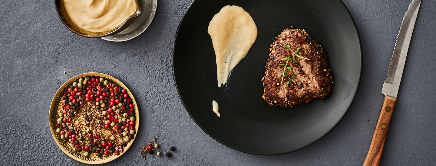 Kypsäksi paistettu pihvi mustalla pyöreällä lautasella. Pihvin viereen on laitettu tilkka vaaleanruskeaa kastiketta. Lautasen vasemmalla puolella on pieni lautanen, jossa on erilaisia kokonaisia pippureita ja pippurirouhetta. Lautasen oikealla puolella on pihviveitsi.