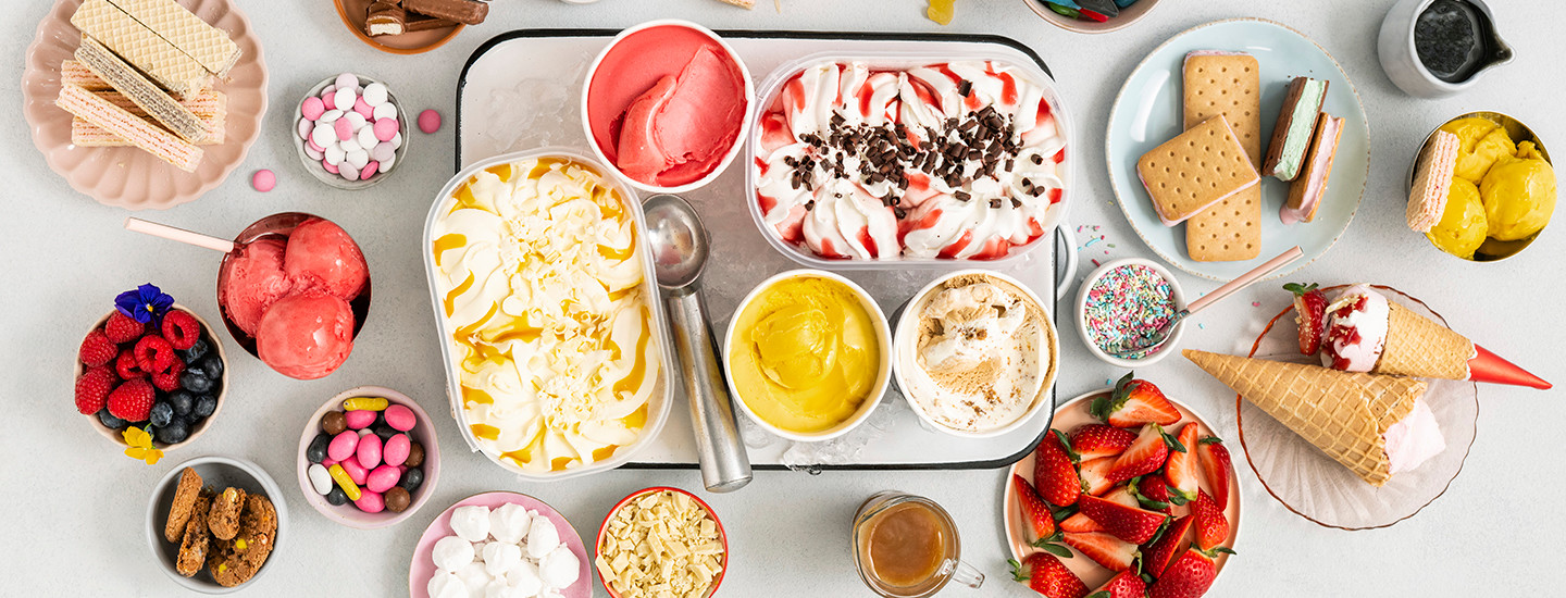Erilaisia jäätelöpakkauksia ja yksittäisiä jäätelötuutteja pöydällä ja tarjottimilla.