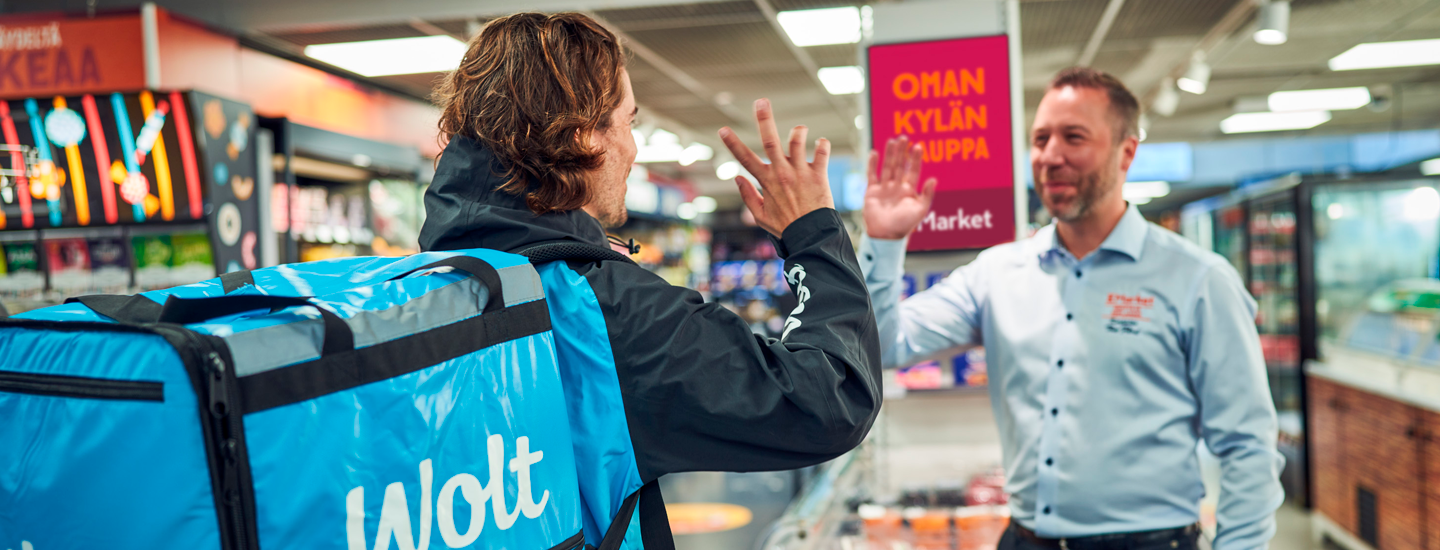 Kaksi miespuolista henkilöä tapaamassa toisensa K-kaupassa ja ovat moikkaamassa toisiaan. Etummainen mies on selkä katsojaan päin, hänen selässä on sininen Wolt-tilauslaatikko. Taaimmaisella miehellä on sinertävä K-Market-kauluspaita päällä.