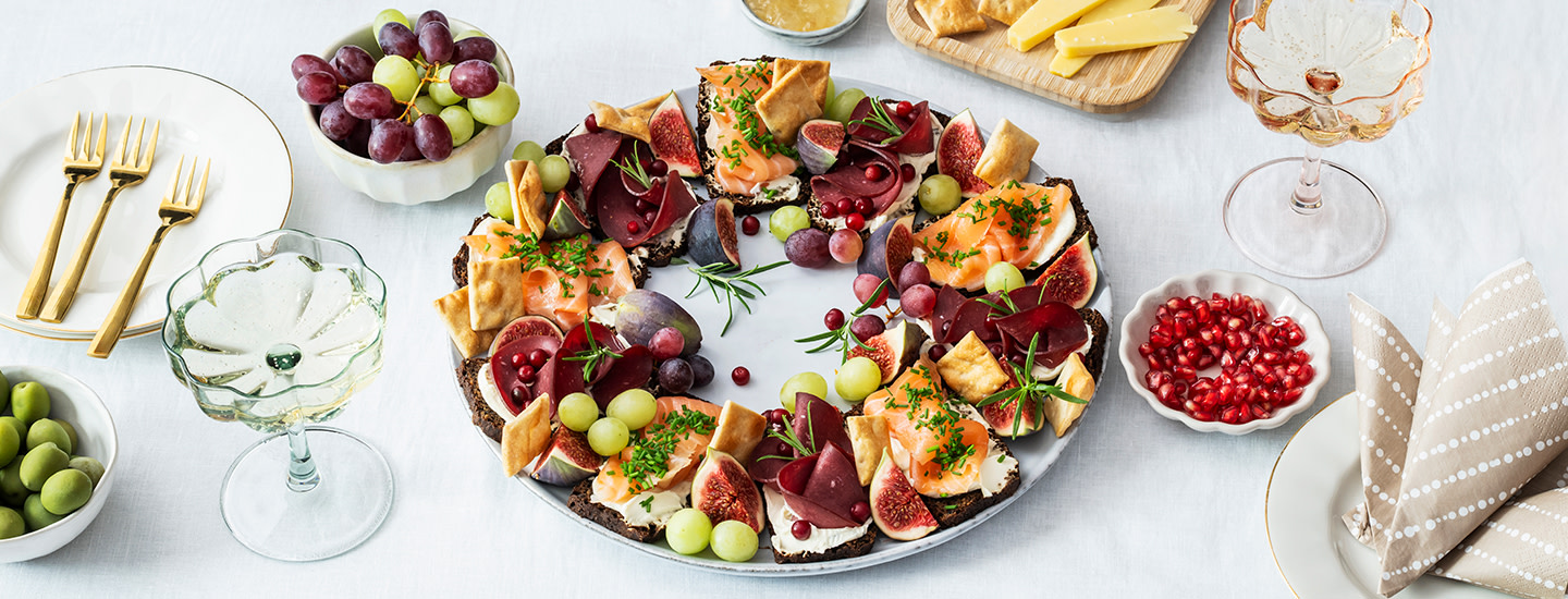 Pieniä leipäpaloja pyöreällä lautasella aseteltuna ympyräksi. Puolet leivistä on päällystetty juustolla ja hillolla, puolet kylmäsavulohella ja granaatinomenansiemenillä. Leipien väliin on aseteltu tuoretta rosmariinia. K-Ruoka alkupalaresepti.