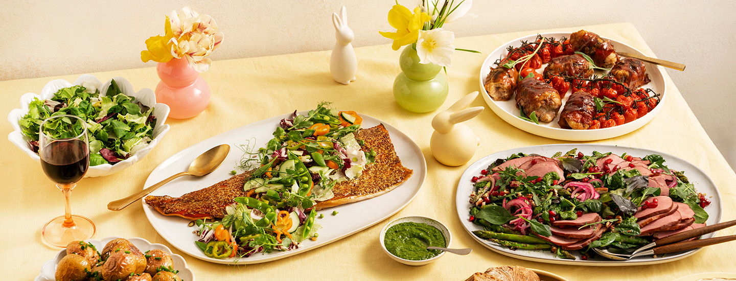 Pöydällä on erilaisia pääsiäisruokia tarjoiluastioissa. Yhdessä astiassa on paistettu lohifilee, jonka päälle on koristeellisesti aseteltu salaattia. Toisessa astiassa on kanasaltimboccaa, paahdettuja kanan paistileikkeitä käärittynä parmankinkkuun. Kolmannessa on vihersalaattia ja karitsanpaistileikkeitä. K-Ruoka pääsiäismenu.