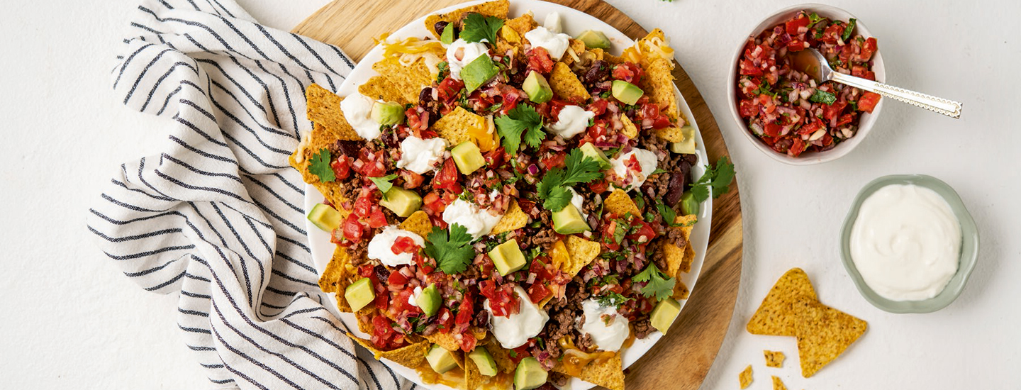 Lautasella nacho-annos, jossa on nachoja, avokadoa, jauhelihaa sekä salsaa. Lautanen on puualustan päällä vaalealla alustalla. K-Ruoan reseptikuva.