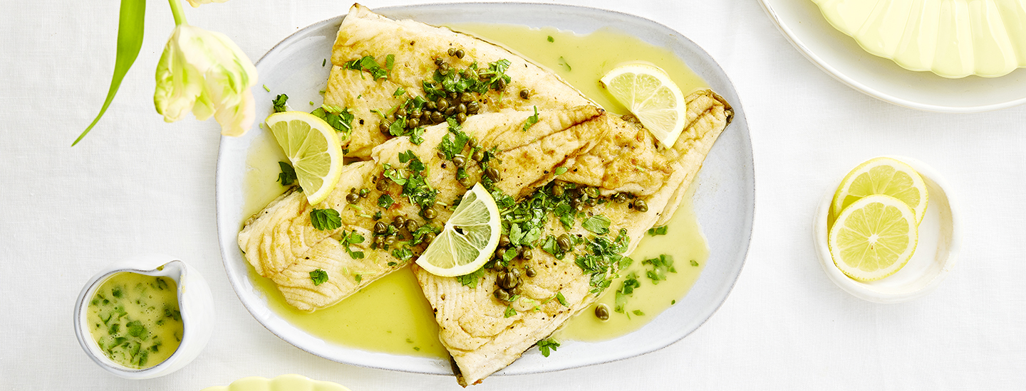 Sitruunavoisiikaa valkoisella ovaalilla tarjottimella. Siianpuolikkaita on tarjottimella kolme. Siikojen päälle on ripoteltu kapriksia ja hienonnettua persiljaa. Tarjottimella on myös sitruunavoikastiketta sekä sitruunanviipaleita. K-Ruoka kalaresepti.