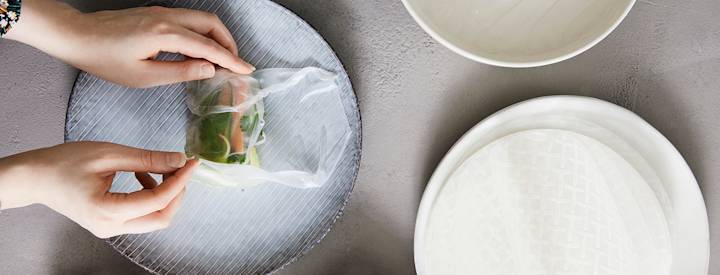 Sinisellä lautasella on kasteltu riisipaperi, jonka keskelle on aseteltu erilaisia täytteitä, kädet ova taittelemassa riisipaperia täytteiden päälle.