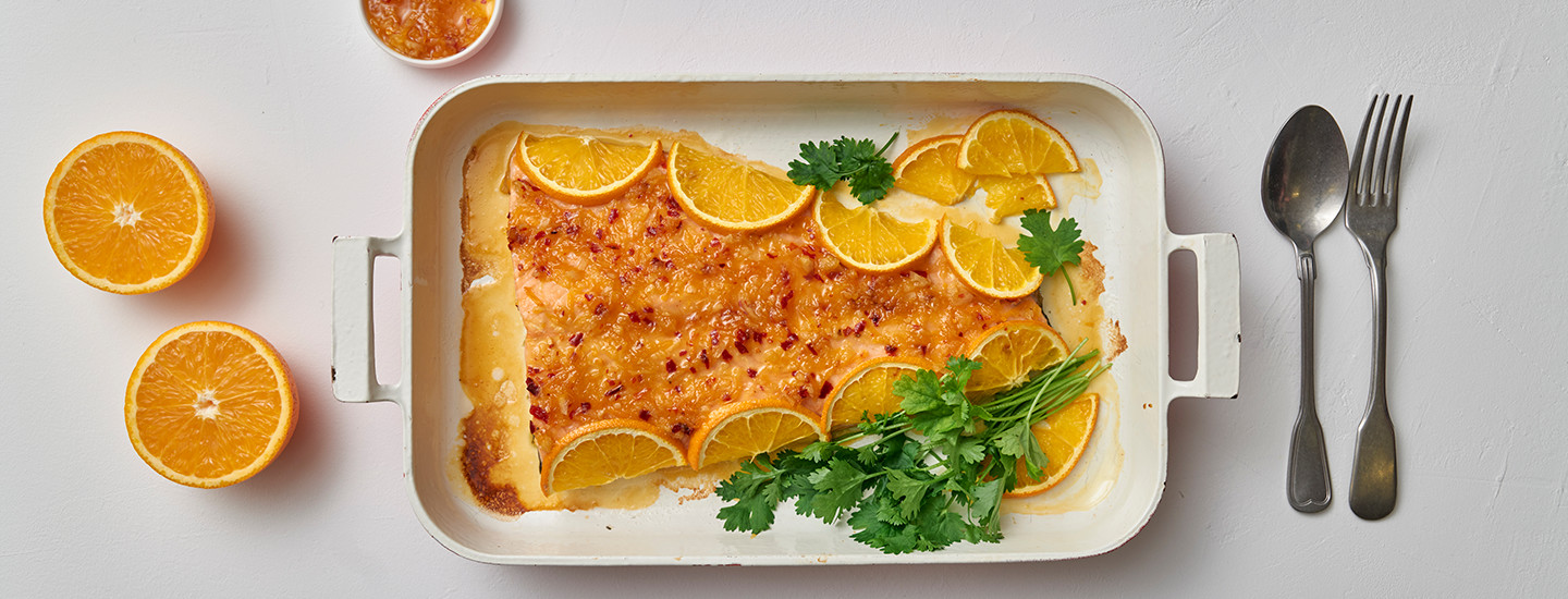Appelsiiniuunilohi valkoisessa uunivuoassa. Lohifileen päälle on aseteltu appelsiinisiivuja ja tuoretta persiljaa. Uunivuoan vasemmalla puolella pöydällä on appelsiininpuolikkaita, oikealla puolella lusikka ja haarukka. K-Ruoka arkiruokaresepti.