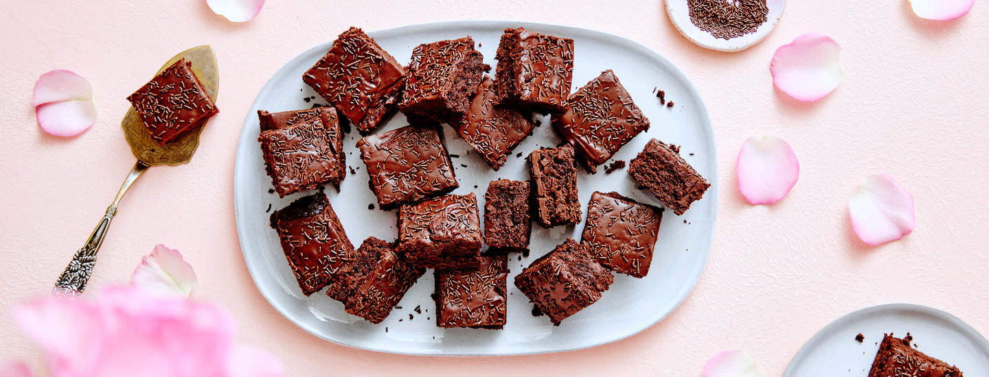 Vegaanisia browniepaloja valkoisella tarjottimella. Tarjottimen ympärillä on vaaleanpunaisia ruusunterälehtiä. K-Ruoka leivontaresepti.