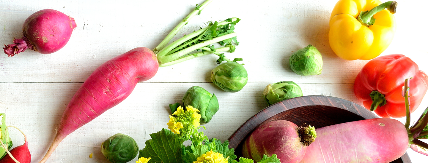 Erilaisia tuoreita kasviksia valkoisella pöydällä. Pöydällä on retiisejä, paprikoita ja ruusukaaleja.