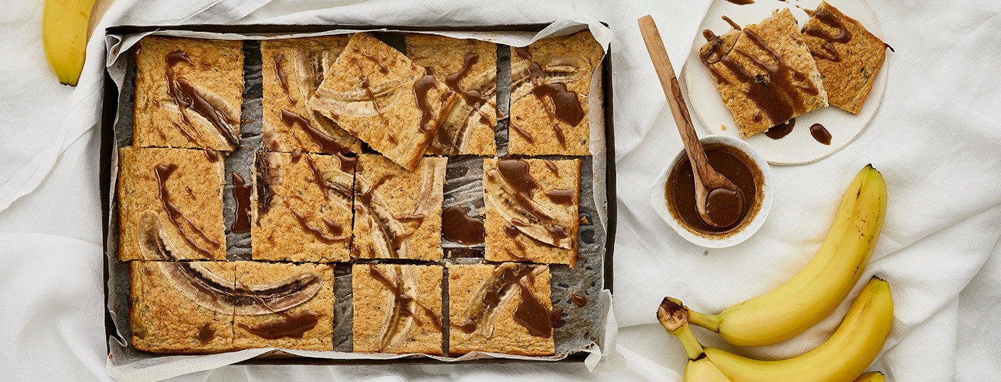 Valkoisella liinalla peitetyllä pöydällä on pellillinen pannukakkua, jonka päällä on kokonaisena viipaloituja banaaneja. Pannukakun päälle on kaadettu pieni määrä ruskeaa tahinikastiketta. K-Ruoka leivontaresepti.