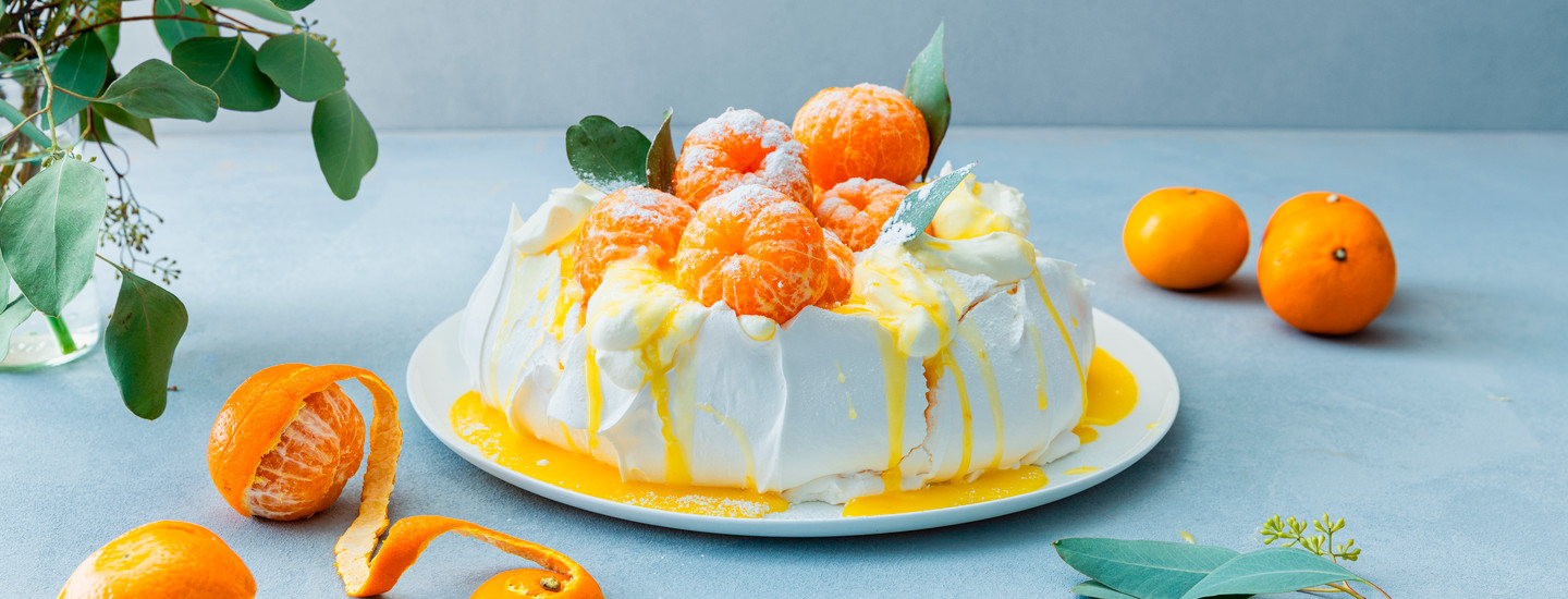 Klementiinipavlova valkoisella pyöreällä lautasella. Pavlova päälle on aseteltu kokonaisia kuorittuja klementiinejä, joiden päälle on ripoteltu tomusokeria. Lisäksi pavlovan päälle on kaadettu klementiinicurdia. K-Ruoka jälkiruokaresepti.