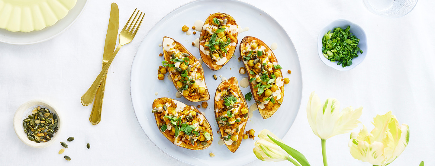 Täytettyjä bataatinpuolikkaita valkoisella pyöreällä lautasella. Bataatit on täytetty kikherne-linssitäytteellä ja päälle on valutettu vaaleaa tahinikastiketta. K-Ruoka arkiruokaresepti.