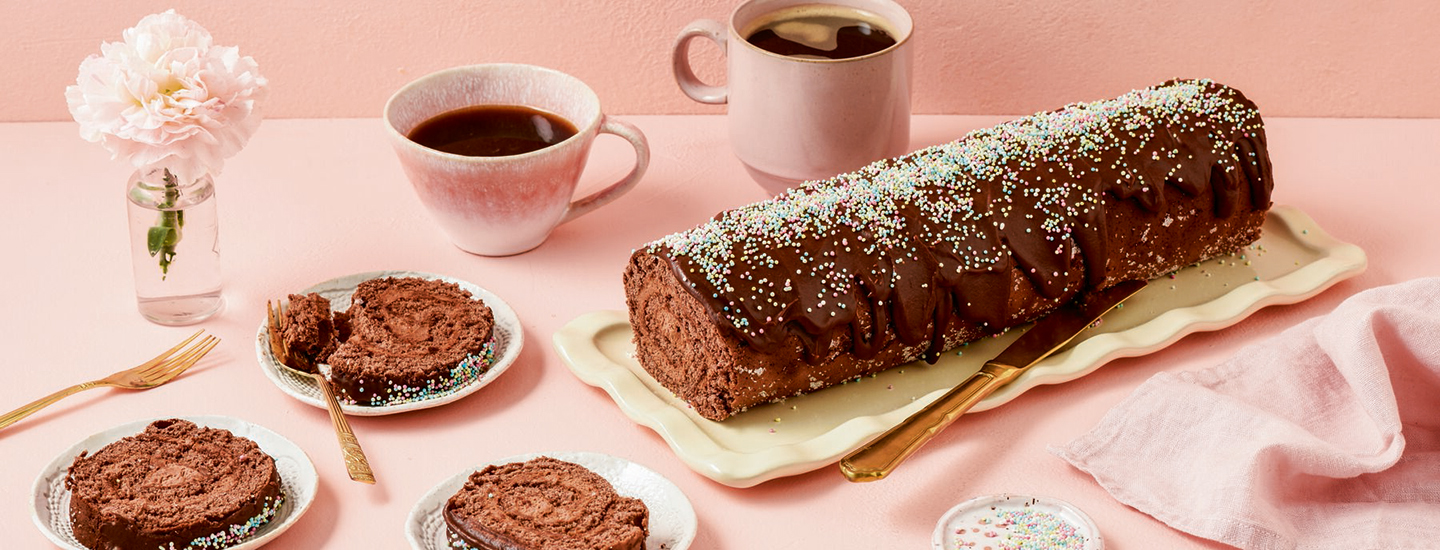 Mokkapalakääretorttu vaaleanpunaisella alustalla. Ympärillä kääretortun paloja ja kahvia. K-Ruoan reseptikuva.