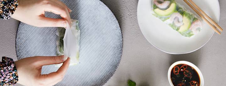 Sinisellä lautasella on kasteltu riisipaperi, jonka keskelle on aseteltu erilaisia täytteitä, kädet ovat taittelemassa riisipaperia täytteiden päälle.