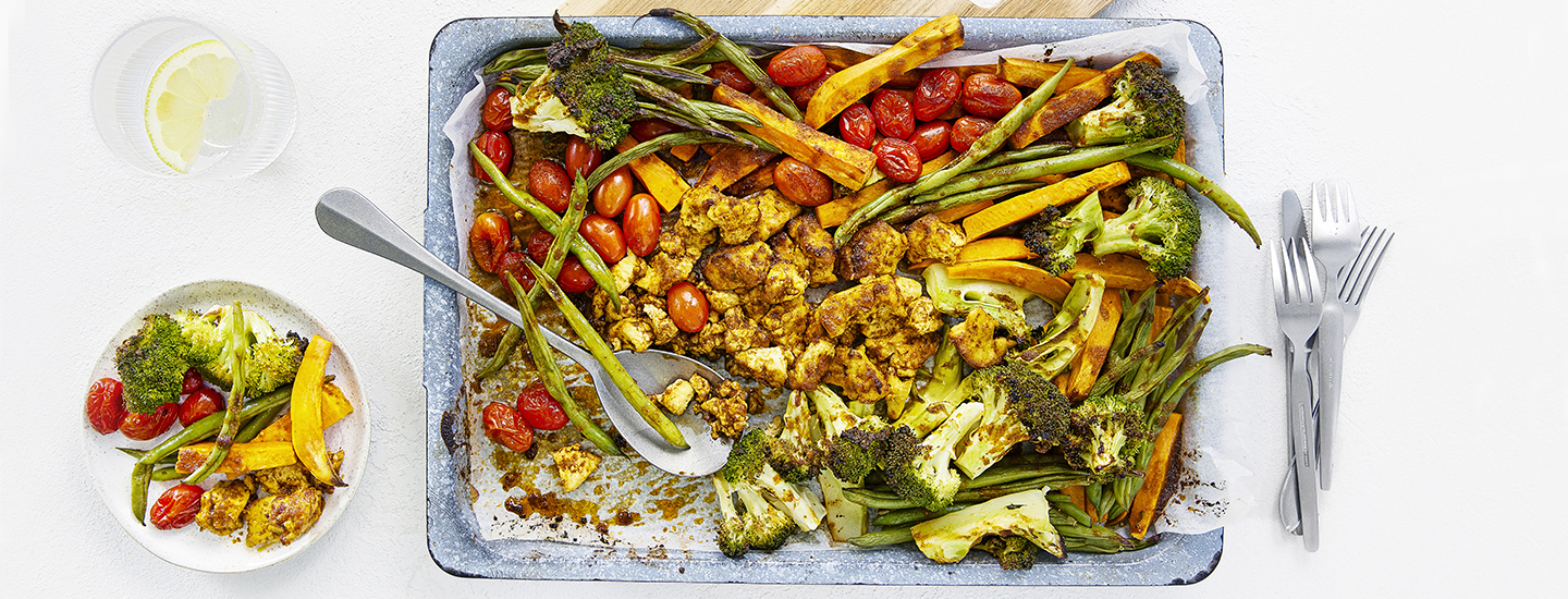 Tofu-tacopelti metallisella pellillä. Pellillä on maustettua tofua, miniluumutomaatteja, parsakaalia, bataattia ja vihreitä papuja. Pellin molemmin puolin on haarukoita, veitsiä, lasillinen vettä ja lautanen, jossa on tofu-tacopeltiruokaa. K-Ruoka vegaaninen arkiruokaresepti.