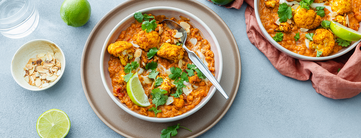 Kasviscurrya valkoisella syvällä lautasella. Ruoan päälle on ripoteltu tuoretta korianteria ja lisätty limesiivu. Valkoinen syvä lautanen on isomman ruskean matalan lautasen päällä. K-Ruoka kasvisruokaresepti.