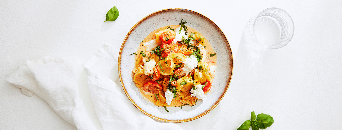 Tortellineja tomaattisessa kermakastikkeessa syvällä lautasella. Lautasella on tortellinien lisäksi mozzarellajuustoa ja tuoreita yrttejä. K-Ruoka pastaresepti.