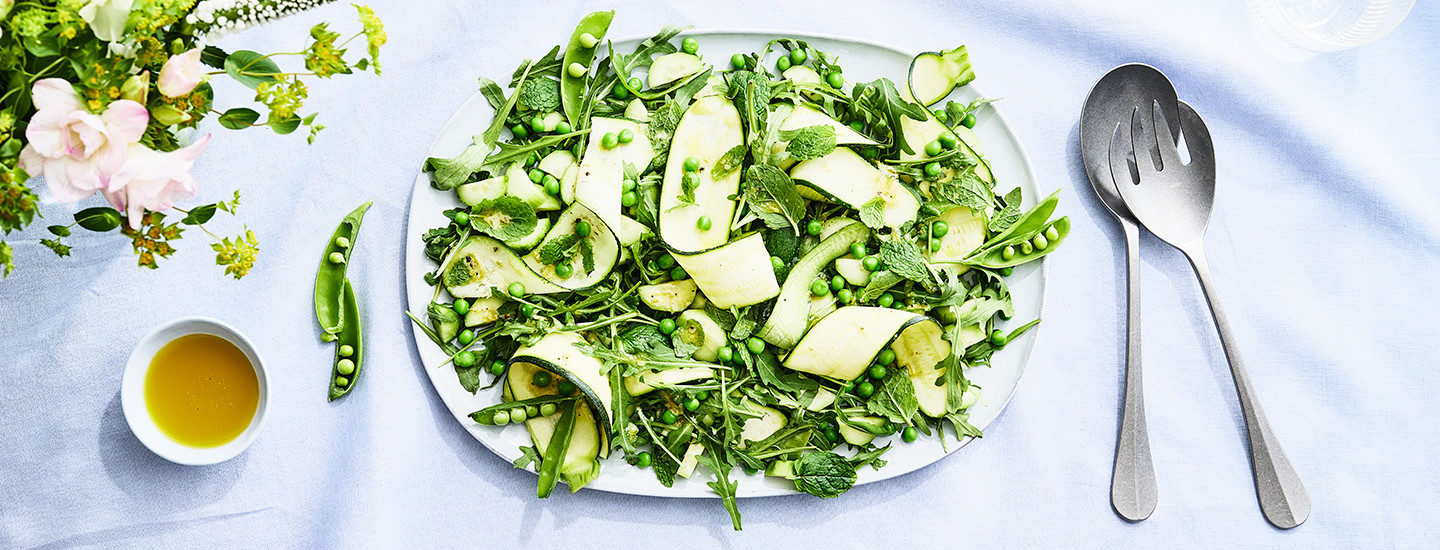 Vihreä hernesalaatti valkoisella tarjottimella, vieressä hopeiset salaattiottimet.