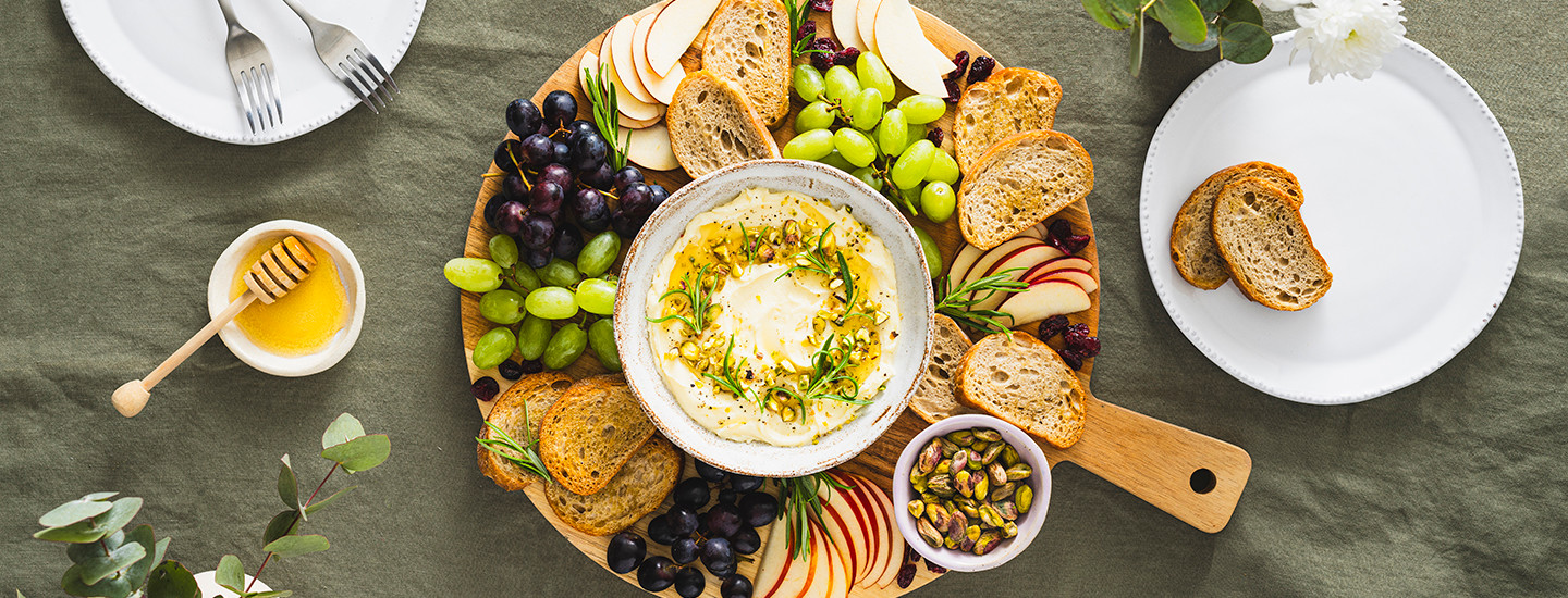 Vatkattu brie pienessä kulhossa puisen pyöreän tarjottimen päällä. Kulhon ympärille on aseteltu tummia ja vaaleita viinirypäleitä, omenaviipaleita ja siivuja vaaleasta leivästä. K-Ruoka naposteluresepti.