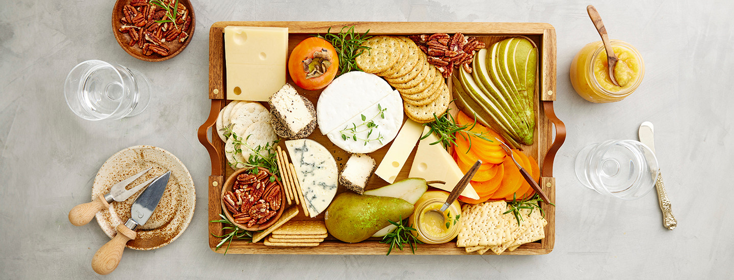 Jouluinen juustotarjotin vaaleanharmaalla pöydällä. Tarjotin on nelikulmion muotoinen ja puinen. Tarjottimella on erilaisia juustoja, keksejä, hedelmiä ja pähkinöitä. K-Ruoka alkupalaresepti.