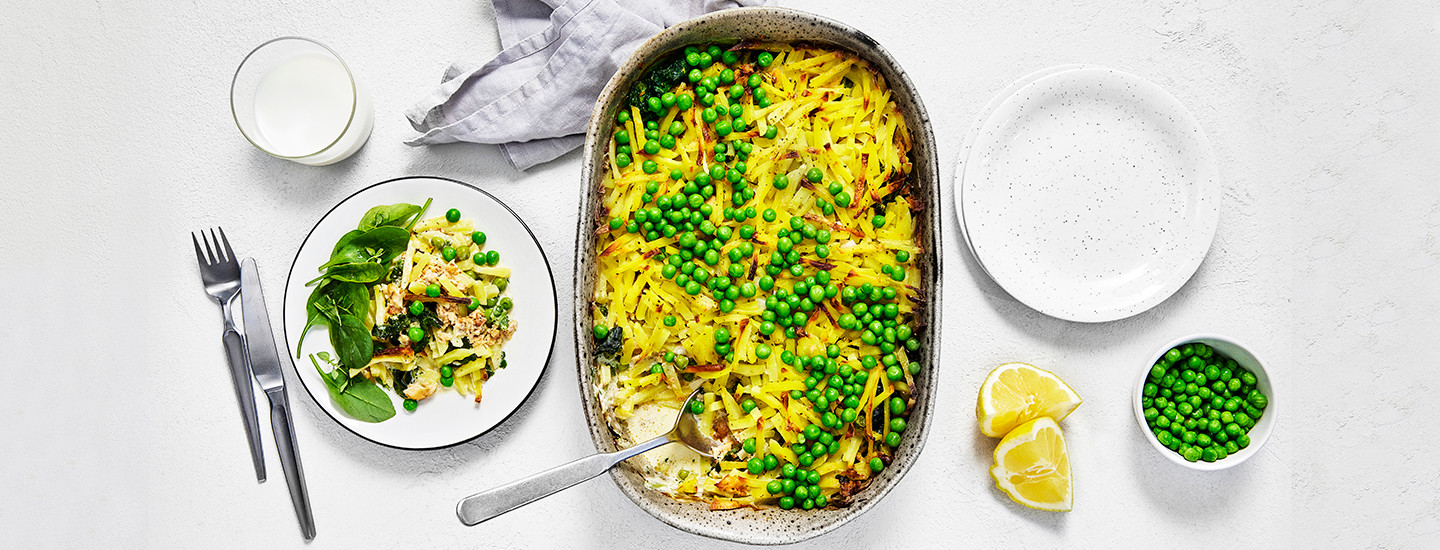 Kalakiusausta harmaassa uunivuoassa. Kalakiusauksen päälle on ripoteltu herneitä. Uunivuoan vasemmalla puolella on lautanen, jossa on annos kalakiusausta ja tuoreita pinaatinlehtiä. Oikealla puolella on kaksi sitruunaviipaletta ja pieni kuppi herneitä. K-Ruoka arkiruokaresepti.