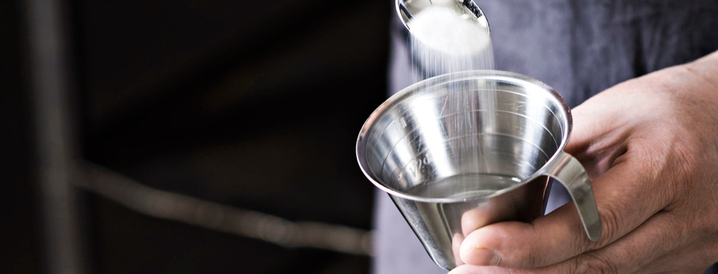 Kädessä on desilitran mitta, johon kaadetaan teelusikasta sokeria.