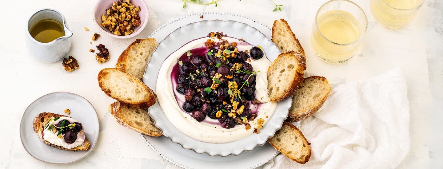 Vaahdotettua ricottajuustoa ja paahdettuja viinirypäleitä lautasella. Lautasen ympärille on aseteltu patonkiviipaleita. K-Ruoka alkupalaresepti.