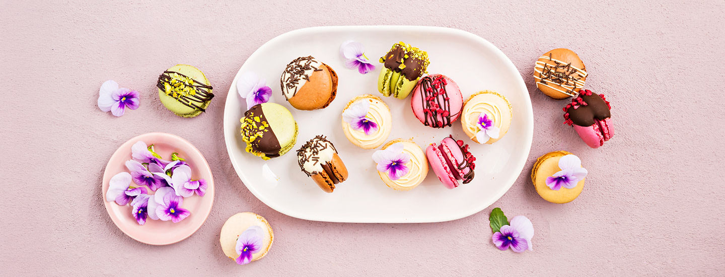 Macaronseja valkoisella tarjoilulautasella. Macaronsit on koristeltu suklaalla, rouhitulla pistaasipähkinällä, strösseleillä, syötävillä kukilla ja vadelmilla. K-Ruoka jälkiruokaresepti.