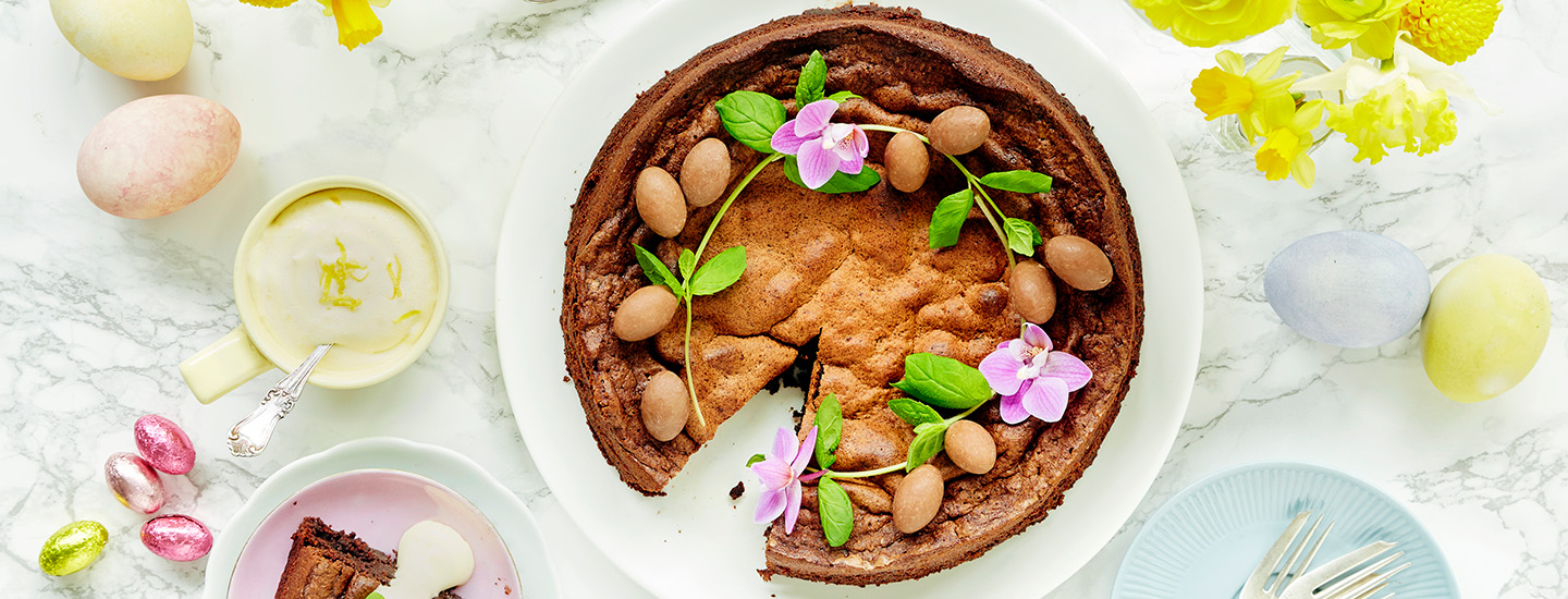 Mämmimudcake valkoisella tarjoilulautasella. Kakun päälle on lisätty suklaamunia, tuoreita mintunoksia ja syötäviä kukkia. K-Ruoka pääsiäisresepti.