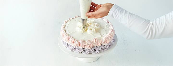 Täytekakku, jonka päälle on levitetty kermavaahtoa. Kakun sivuille on tehty vaaleanliiloja ja vaaleanpunaisia ruusupursotuksia. Kakun päälle ollaan tekemässä valkoisia ruusupursotuksia.