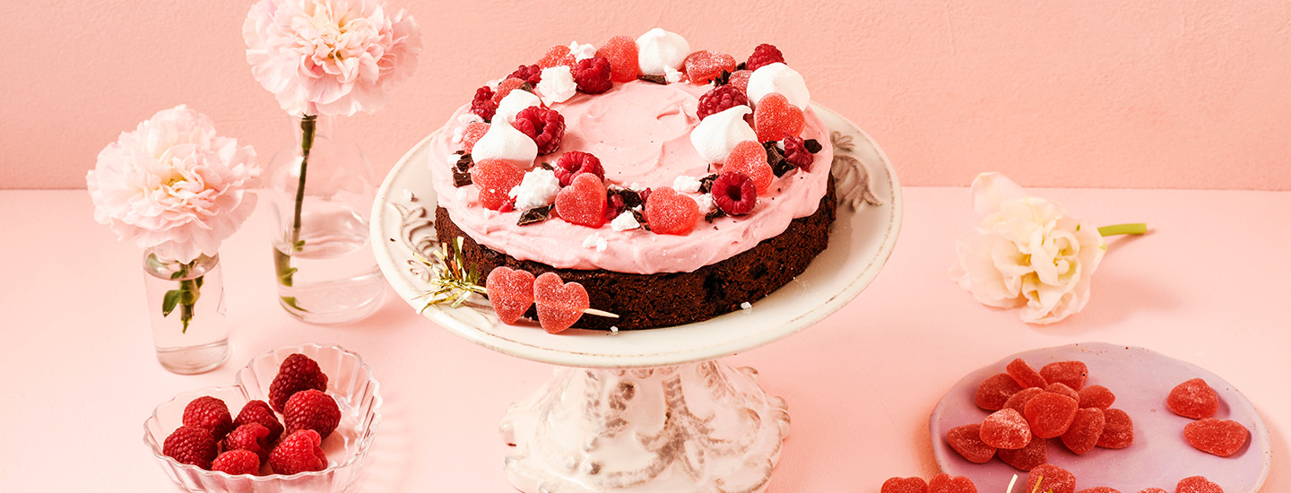 Kaakaosta tehty tumma kakkupohja kakkutarjottimella. Kakku on kuorrutettu vaaleanpunaisella kuorrutteella ja koristeltu sydänmarmeladeilla, marengeilla, marjoilla ja suklaarouheella. K-Ruoka kakkuresepti.