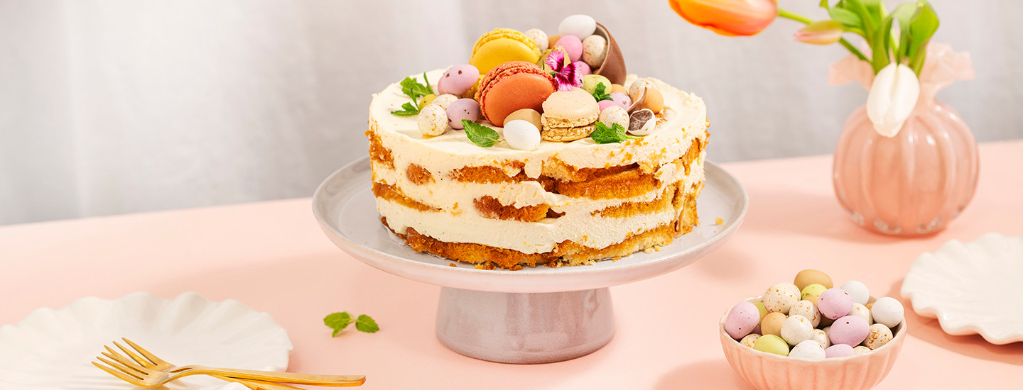 Pääsiäiskakku valkoisella kakkutarjottimella. Kakku on naked cake -tyylinen, se on koottu valmiin sitruunakuivakakun paloista ja mascarpone-kermavaahdosta. Kakun päälle on lisätty suklaamunia, macaronseja ja tuoreita mintunlehtiä. K-Ruoka kakkuresepti.