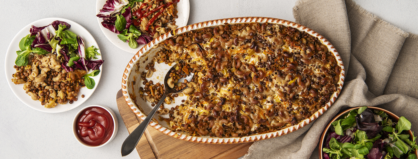 Linssimakaronilaatikkoa ovaalinmuotoisessa uunivuoassa. Uunivuoan vasemmalla puolella on pyöreä lautanen, johon on laitettu linssimakaronilaatikkoa ja salaattia. Lautasen vieressä on pieni kulho, jossa on ketsuppia. K-Ruoka arkiruokaresepti.