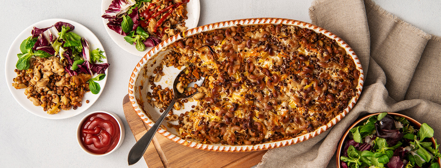 Linssimakaronilaatikkoa ovaalinmuotoisessa uunivuoassa. Uunivuoan vasemmalla puolella on pyöreä lautanen, johon on laitettu linssimakaronilaatikkoa ja salaattia. Lautasen vieressä on pieni kulho, jossa on ketsuppia. K-Ruoka arkiruokaresepti.