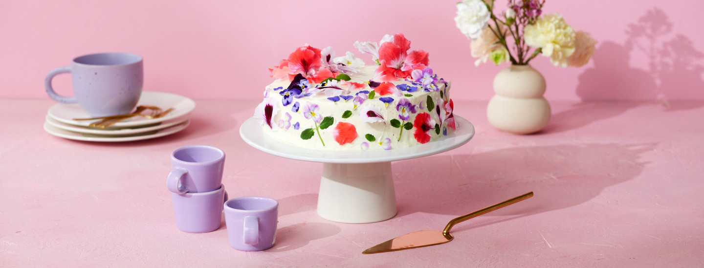 Kermavaahdolla ja syötävillä kukilla koristeltu gluteeniton täytekakku, joka on valkoisella kakkutarjoiluastialla vaaleanpunaisen pöydän päällä.