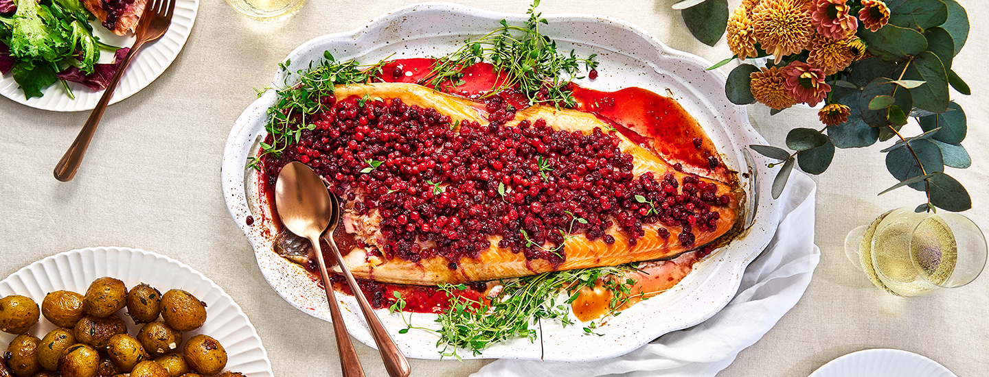 Puolukoilla höystetty lohi tarjoiluastialla. Pöydällä muita ruokatarjoiluja. K-Ruoan reseptikuva.