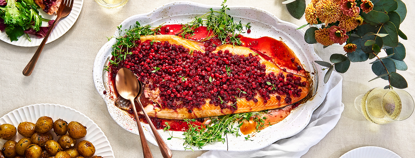 Puolukoilla höystetty lohi tarjoiluastialla. Pöydällä muita ruokatarjoiluja. K-Ruoan reseptikuva.