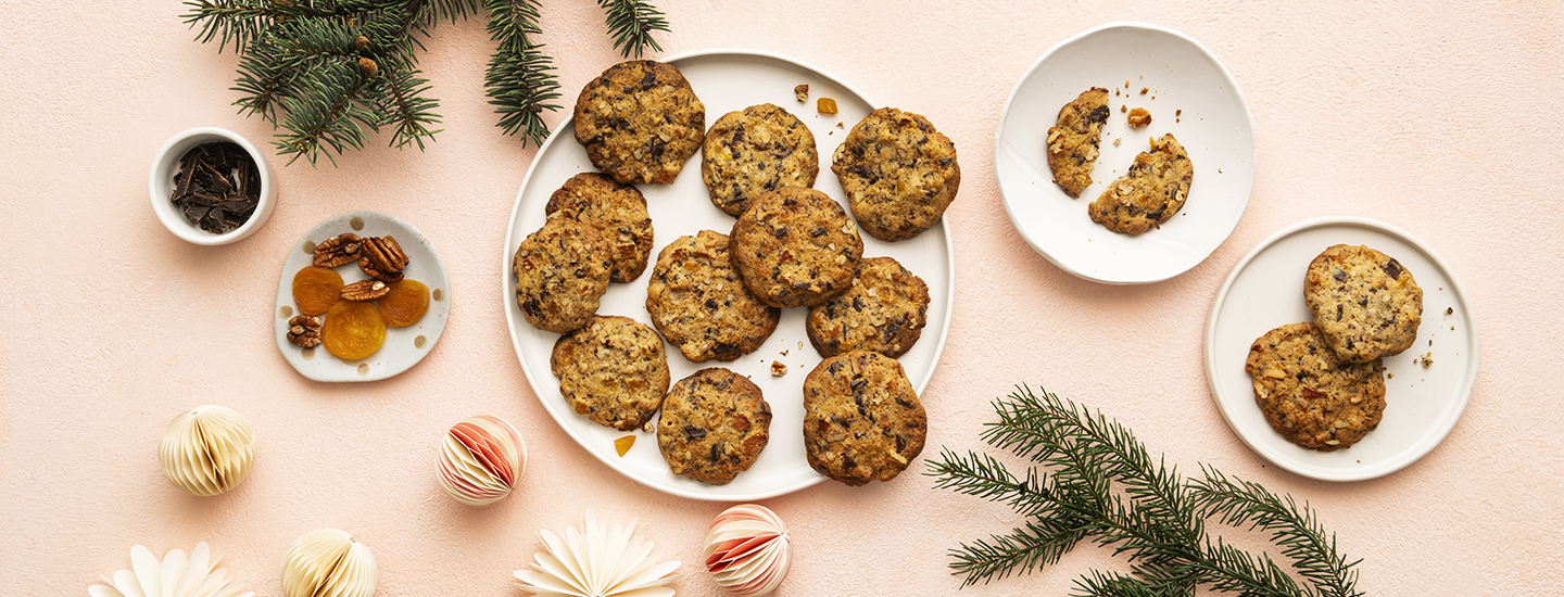 Joulukeksejä valkoisella tarjoilulautasella. Kekseissä on suklaahippuja, pähkinöitä ja kuivattua aprikoosia. Tarjoilulautasen ympärillä on jouluisia koristeita: joulupalloja ja kuusenoksia. K-Ruoka keksiresepti.