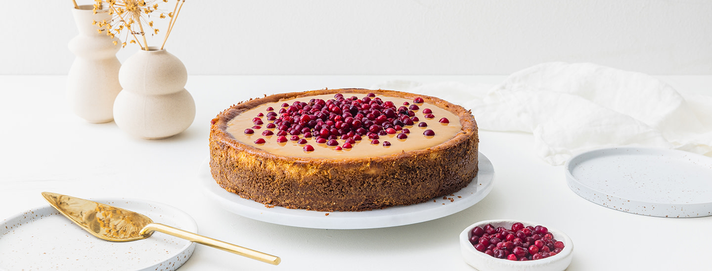 Puolukka-juustokakku valkoisella pyöreällä kakkulautasella. Juustokakun päällä on kinuskikerros ja puolukoita. K-Ruoka juustokakkuresepti.