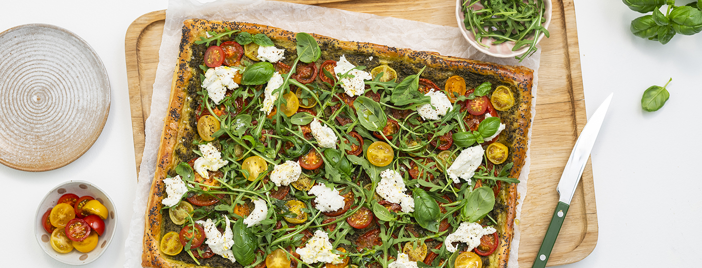 Suorakulmion muodossa oleva tomaatti-pestopiirakka leivinpaperin ja puisen leikkuulaudan päällä. Piirakan päällä on pestokastiketta, punaisia ja keltaisia minitomaatin puolikkaita, revittyä mozzarellaa ja tuoretta rucolaa sekä basilikaa. K-Ruoka piirakkaresepti.