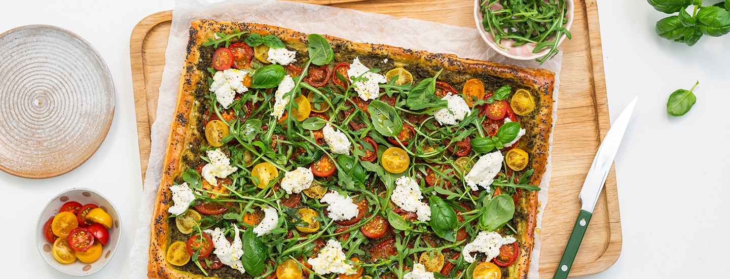 Suorakulmion muodossa oleva tomaatti-pestopiirakka leivinpaperin ja puisen leikkuulaudan päällä. Piirakan päällä on pestokastiketta, punaisia ja keltaisia minitomaatin puolikkaita, revittyä mozzarellaa ja tuoretta rucolaa sekä basilikaa. K-Ruoka piirakkaresepti.