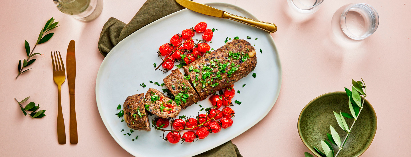 Vaaleanpunaisella taustalla katettu lautanen, jossa täytettyä lihamureketta sekä tomaatteja vierellä. Lautasen vierellä vihreä kulho ja lautasen alla vihreä liina. Oikealle puolelle on katettu kultaiset aterimet. K-Ruoka jauheliharesepti.