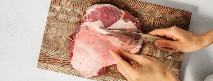 Karitsan valmistuksen ensimmäinen vaihe: karitsanpaistin sulatus ja siistiminen. Puisella leikkuulaudalla on raaka karitsanpaisti, jota leikataan veitsellä.