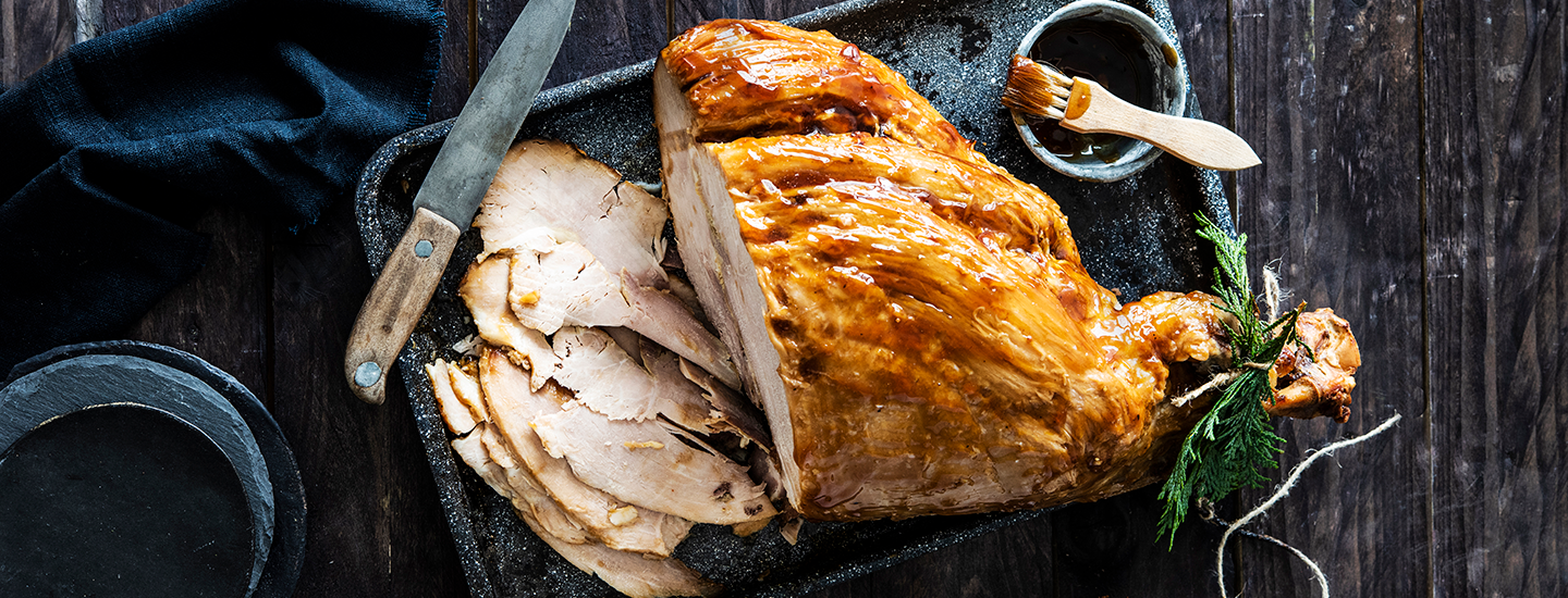 Glaseerattu joulukinkku mustalla tarjottimella. Kinkusta on leikattu siivuja, jotka ovat kinkun vieressä tarjottimella. Kinkun vieressä on myös pieni kulho glaseerauskastiketta. K-Ruoka jouluresepti.