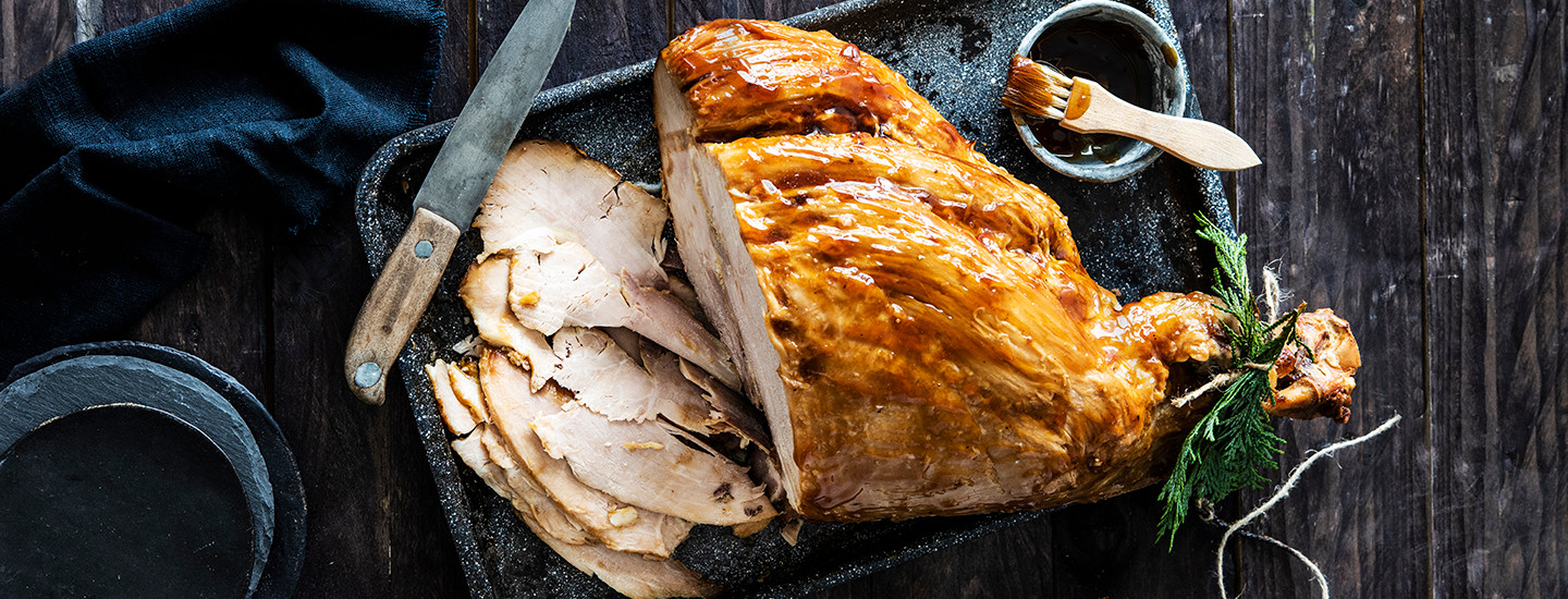 Glaseerattu joulukinkku mustalla tarjottimella. Kinkusta on leikattu siivuja, jotka ovat kinkun vieressä tarjottimella. Kinkun vieressä on myös pieni kulho glaseerauskastiketta. K-Ruoka jouluresepti.