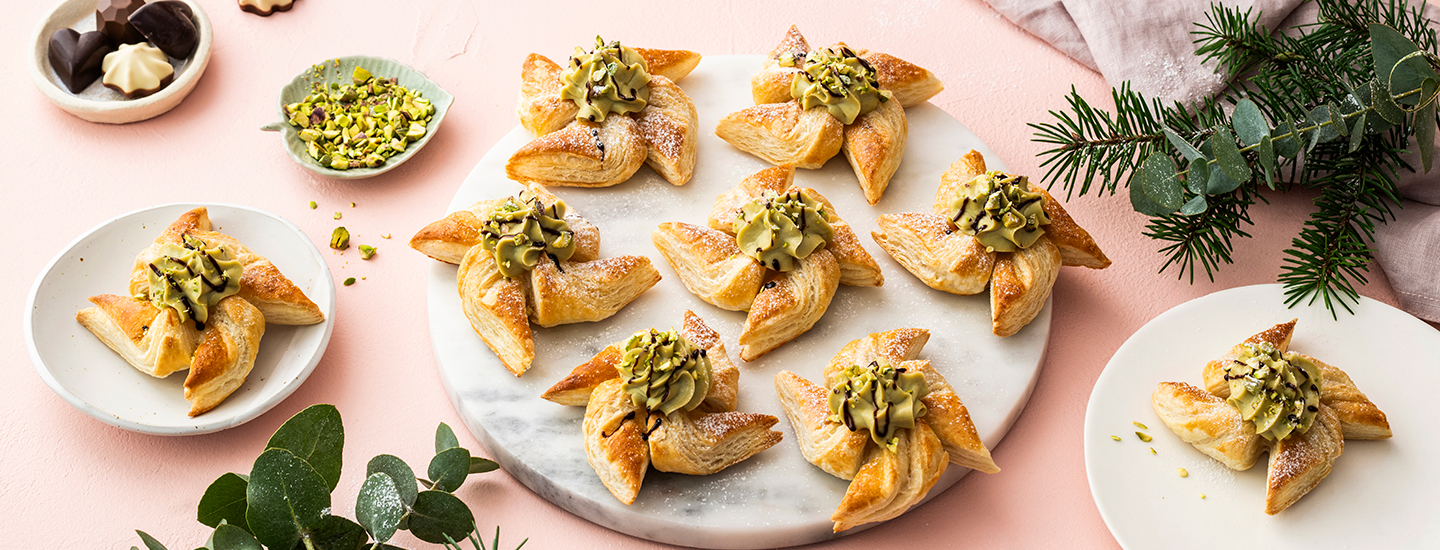 Joulutorttuja pistaasitahnatäytteellä pyöreällä tarjoilulautasella. Torttujen päälle on lisätty suklaakastiketta ja pistaasimurua. K-Ruoka jouluresepti.