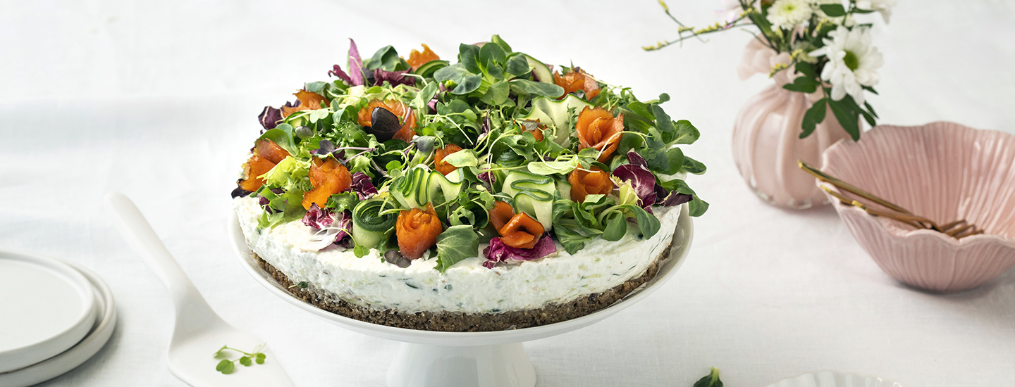 Tsatsiki-lohivoileipäkakku valkoisella tarjoilulautasella. Voileipäkakku on pyöreä ja juustokakkumaiseen tyyliin valmistettu, siinä on leipäpohja ja tsatsikitäyte. Voileipäkakun päällä on salaattia, kylmäsavulohta ja kurkkua. K-Ruoka voileipäkakkuresepti.