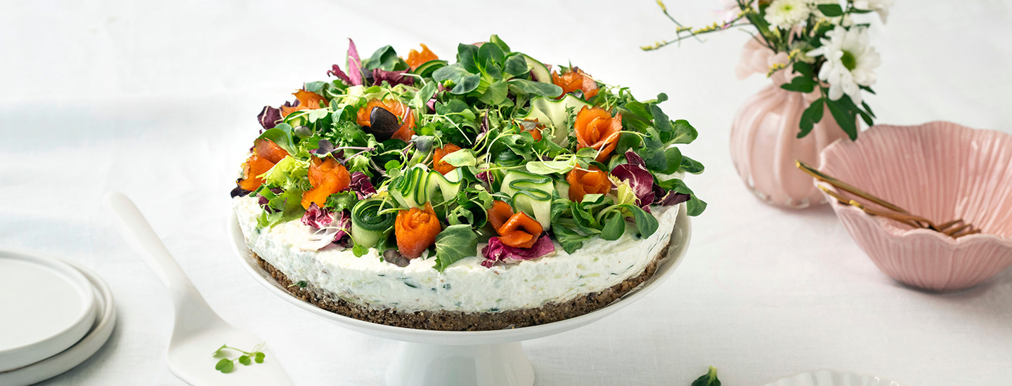 Tsatsiki-lohivoileipäkakku valkoisella tarjoilulautasella. Voileipäkakku on pyöreä ja juustokakkumaiseen tyyliin valmistettu, siinä on leipäpohja ja tsatsikitäyte. Voileipäkakun päällä on salaattia, kylmäsavulohta ja kurkkua. K-Ruoka voileipäkakkuresepti.