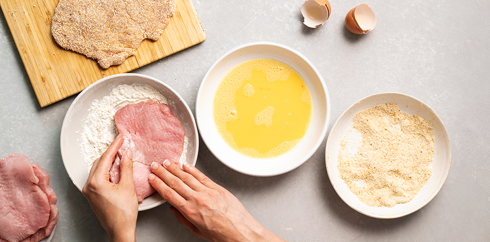 Wieninleikettä ollaan kastamassa kulhossa oleviin vehnäjauhoihin. Vehnäjauhokulhon vieressä on kaksi muuta kulhoa: toisessa on kananmunaa ja toisessa korppujauhoja.