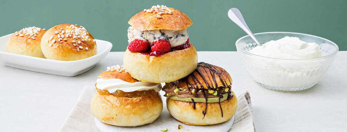 Kolme laskiaispullaa lautasella eri täytteillä. Yhdessä pullassa sisällä on kermavaahtoa ja omenahilloa, toisessa pullassa sisällä on lakritsijäätelä ja vadelmahilloa, kolmannessa pullassa sisällä on suklaamoussea ja pistaasilevitettä. K-Ruoka laskiaispullaresepti.