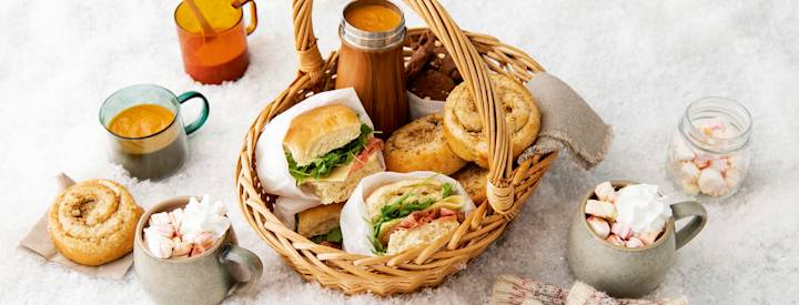 Puisessa korissa retkieväitä. Kori on lumisella alustalla, ympärillä kaakaota, keittoa ja pullia.